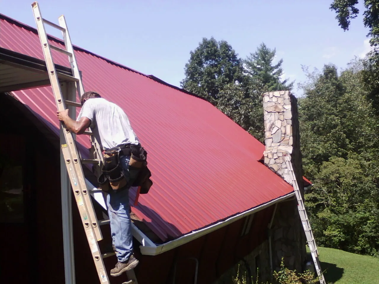 Roofer on a ladder beside a red metal roof and stone chimney — Soffit & Fascia Repair by Roofing Brothers in Powellsville